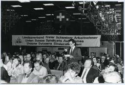 Personen hintereinander an Tischen sitzend; im Hintergrund Banner mit Aufschrift "Landesverband Freier Schweizer Arbeitnehmer / Union Suisse Syndicats Autonomes / Unione Svizzera Sindacatti Liberi"; ein Mann steht vor einem Mikrofon und spricht zum Publikum; 60. Delegiertenversammlung 11. Mai 1985, Hotel Nova-Park Zürich