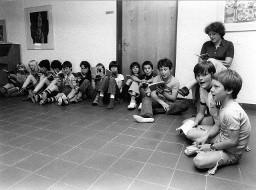 Kinder sitzen am Boden und singen aus einer Liedersammlung - Frau auf dem Stuhl sitzend