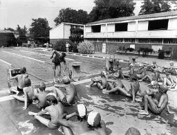 Kinder sitzen vor den Sprungbrettern (Schwimmbad) - Schwimmlehrer Edwin Frey das Kraulschwimmen ausserhalb des Schwimmbads nachahmend, Magliaso 1978