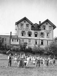 Gebäude mit der Beschriftung: "Gasthaus zur Linde" - Kinder und Erwachsenen auf der Wiese vor dem Gebäude - Sichel und Gabel