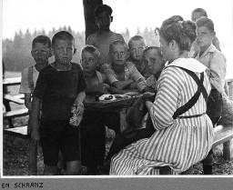En Schranz - Knaben sitzen und stehen im Freien um einenTisch herum - Knaber ein Stück Brot in der Hand haltend - Frau nähend (flickend)