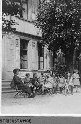 Strickstunde - Mädchen und Frau sitzen auf Stühlen vor dem Ferienkoloniehaus und stricken