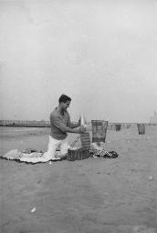 Bob Steffen am Strand auf Picknickdecke, packt Sachen in Picknickkorb; im Hintergrund das Pier, 1961