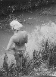 Thea Kuiper, mit Kopftuch, steht an Wasserlauf mit Schilf, 1966