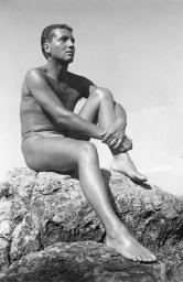 Bob Steffen in Badehose sitzt mit einem Knie angewinkelt auf Felsen und blickt in die Ferne, um 1950