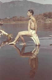 Junger Mann in heller Badehose sitzt auf einem im Wasser liegenden Baumstamm, Spiegelung, um 1945