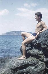 Junger Mann in blauen Badehosen sitzt auf Felsen und schaut aufs Meer, um 1950
