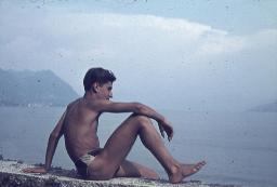 Junger Mann in Badehosen auf Mauer am See, Profilansicht, um 1950