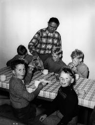 Fünf Kinder sitzen am Tisch (Mahlzeit, vermutlich Frühstück) und ein Mann sie bedienend