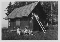 "Weekend Häuschen des Mädchenklubs Fides Zürich", Versammlung mehrerer Kinder bei einem Haus, drei Kinder auf einer Leiter sitzend, zwei Kinder auf Stühlen sitzend und ein Kind in der Wiese sitzend, um 1950