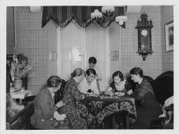 Sechs Personen in einem Raum spielen "Hütchenspiel", fünf Personen sitzend, eine Person stehend, um 1940