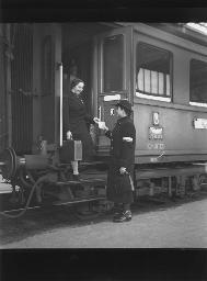 Eine Frau mit Koffer aus einem Zug aussteigend, eine Bahnhofshilfe "Freundinnen junger Mädchen" empfängt sie, um 1920