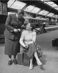 Zwei Frauen miteinander sprechend, eine Frau des Bahnhofhilfwerks stehend und eine Frau auf Koffern sitzend, um 1950