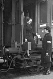 Eine Frau mit Koffer aus einem Zug aussteigend, eine Bahnhofshilfe "Freundinnen junger Mädchen" empfängt sie, um 1950