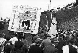 Plakat "An das Schandbrett mit den Heimatschändern", getragen am Fussmarsch von Demonstrationsteilnehmern anlässlich der Kundgebung der Rheinaubewegung am 31.08.1952. Das Plakat zeigt einen Unternehmer mit Zylinderhut im Pranger.