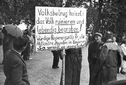 Einzelne Veranstaltungsteilnehmer umringen Plakat "Volksbetrug heisst: das Volk ruinieren und lebendig begraben! Würdige Regierungsräte? als Aktionäre, Magnaten in Konzernen – so verkauft man unsere Freiheit!" Kundgebung der Rheinaubewegung am 31.08.1952