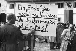 Mann mit handschriftlichem Plakat: "Ein Ende den unsauberen Verträgen: Wahl des Bundesrats durch das Volk", an der Kundgebung der Rheinaubewegung am 31.08.1952. Dessen Vorderseite wird von einer Veranstaltungsteilnehmerin begutachtet.