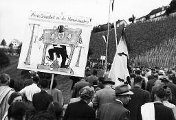 "Auf dem Heimweg" – Plakat "An das Schandbrett mit den Heimatschändern", getragen am Fussmarsch von Demonstrationsteilnehmern anlässlich der Kundgebung der Rheinaubewegung, 31.08.1952. Das Plakat zeigt einen Unternehmer mit Zylinderhut im Pranger.