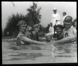 "Zürich, Töchterheim Jakobstrasse", mehrere junge Frauen geben sich im Wasser die Hand, alle mit Badkappe und Badkleider, am Ufer drei weitere Frauen in weisser Kleidung, um 1930